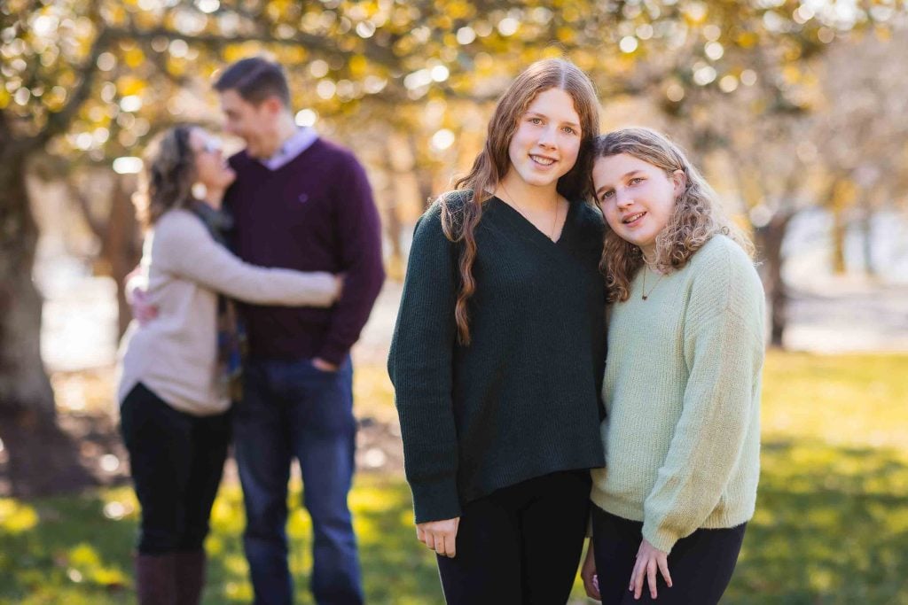 Two young women smiling at the camera in the foreground, with a couple embracing in the blurred background in a sunny park, perfect for family portraits.