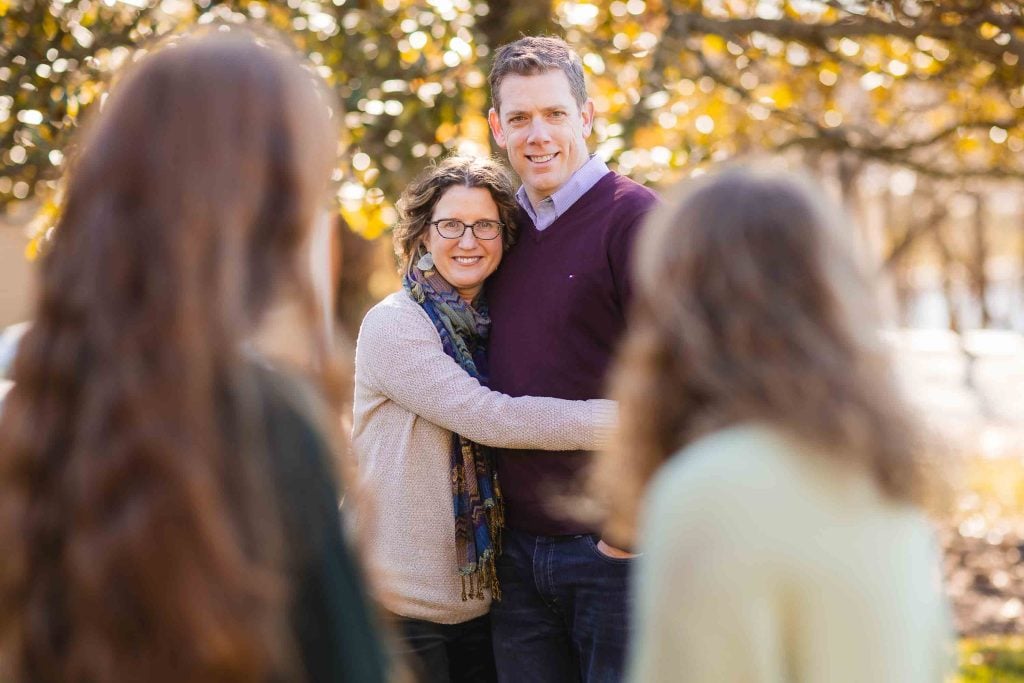 A family portraits of a couple smiling at the camera, embraced outdoors with blurred children in the foreground, set in a warm, autumnal setting with trees.