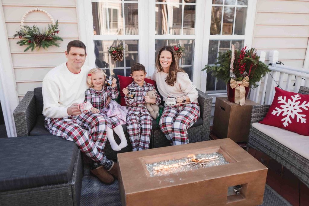 A family of four wearing matching plaid pajamas, enjoying Family Portraits while sitting together with mugs in hand on a porch decorated for Christmas with a fire pit in front.