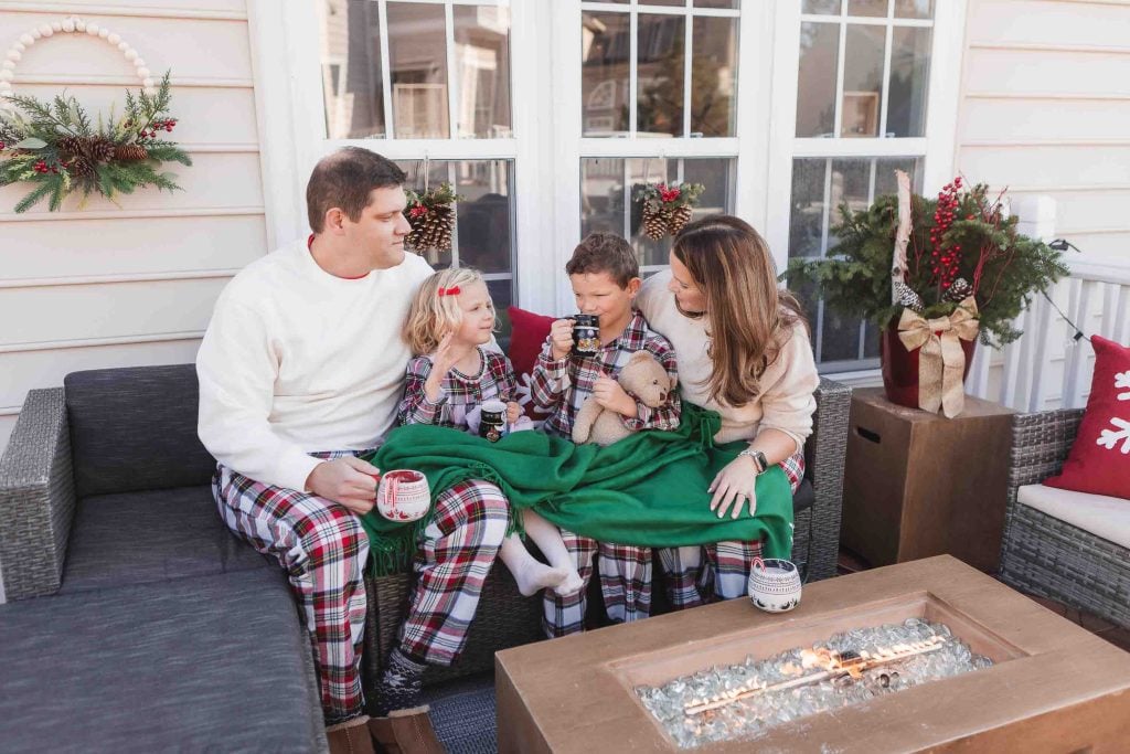 A family of four, dressed in holidaythemed pajamas, enjoying a warm drink together on an outdoor porch decorated for Christmas, perfect for Family Portraits.