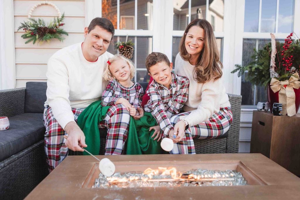 A family of four roasting marshmallows on a fire table in front of a house decorated for Christmas, perfect for Family Portraits.