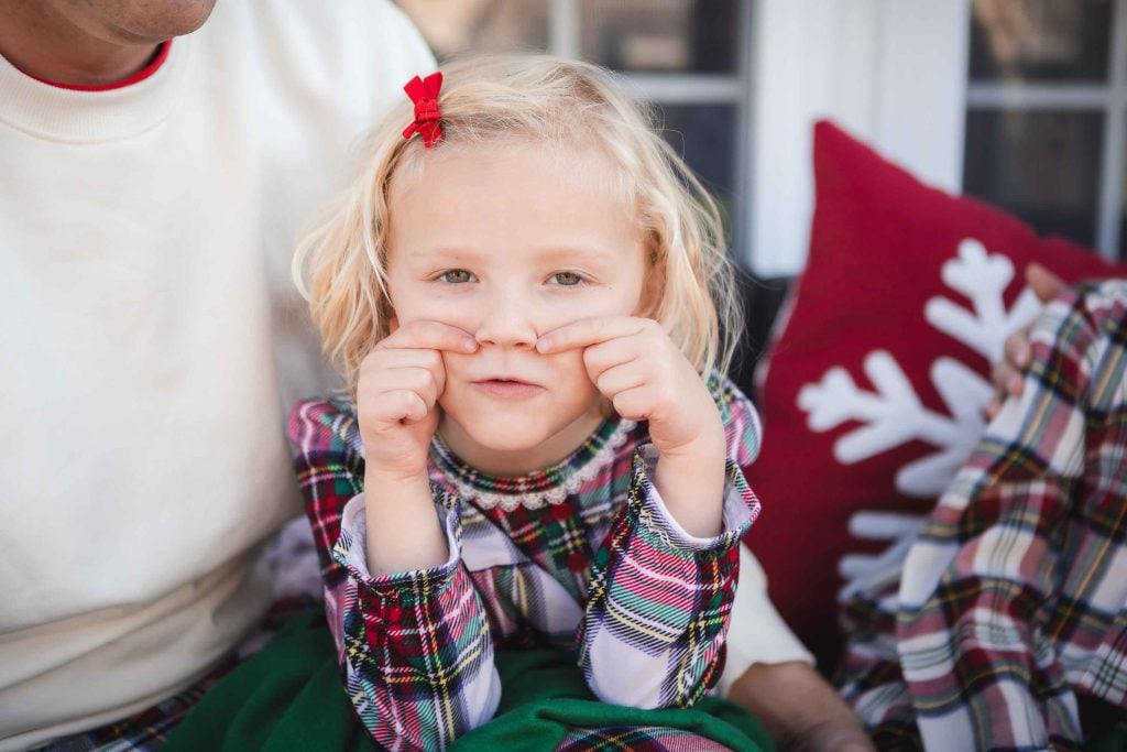 A young girl pulling a funny face by tugging at her nose, wearing festive clothing in a family portrait, with a partial view of an adult beside her.