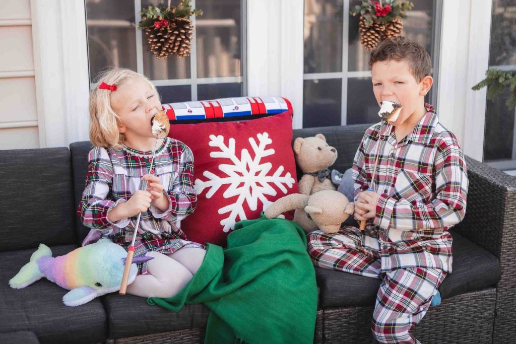 Two children in plaid pajamas sitting on a porch, laughing and eating marshmallows, with a holidaythemed pillow beside them.