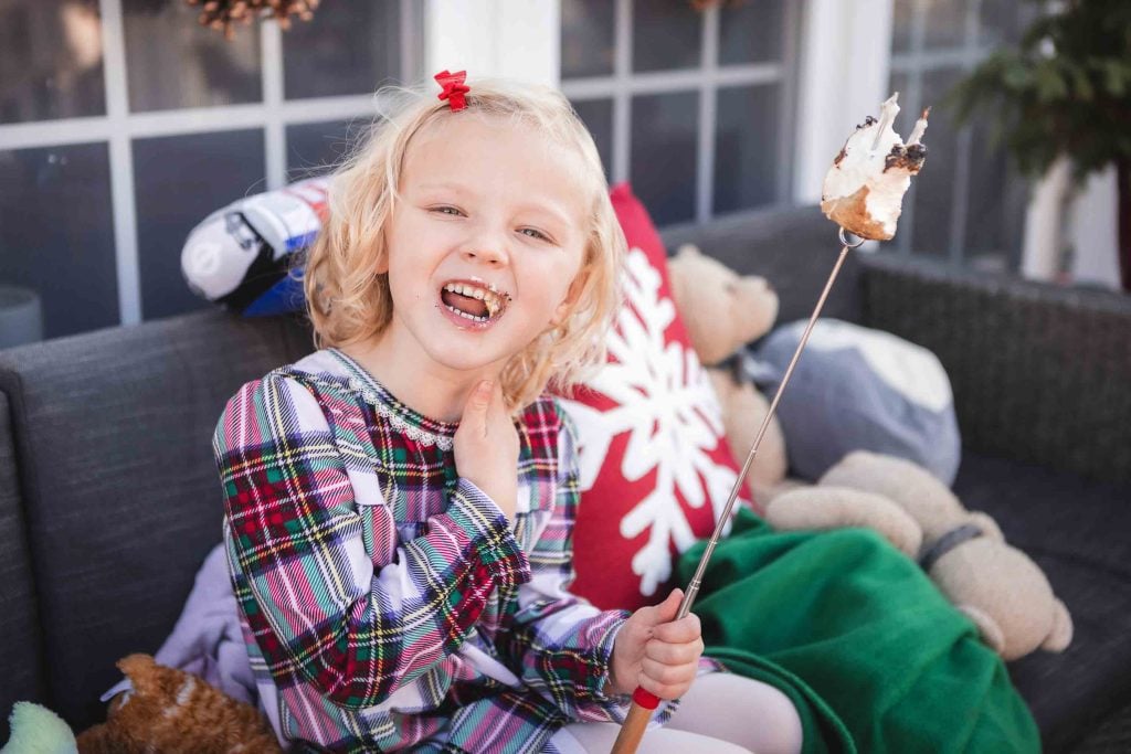 A young girl smiling with marshmallow on her face, holding a stick with a toasted marshmallow, sitting on an outdoor couch with plush toys around during a family portraits session.