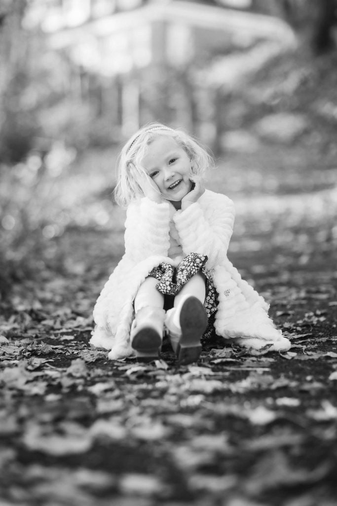 A young girl smiling, sitting on a leafcovered path, wearing a white coat and sparkly shoes, in a black and white family portrait photo.