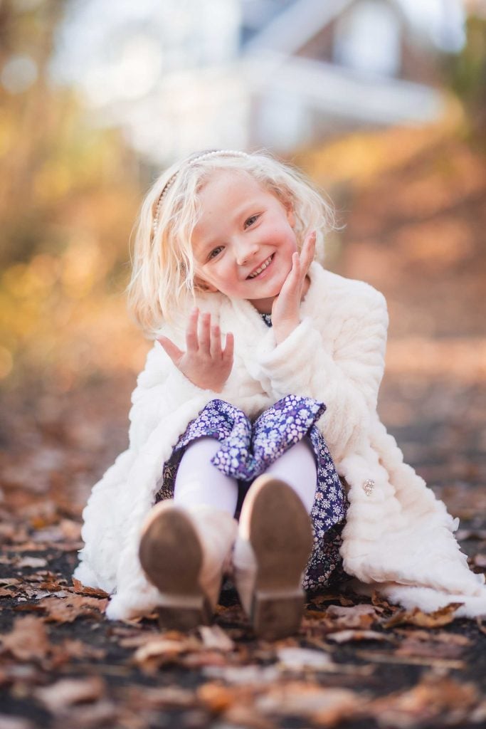 A young girl with blonde hair smiling and waving, sitting on a leafcovered path in a family portrait, wearing a white coat and patterned dress.