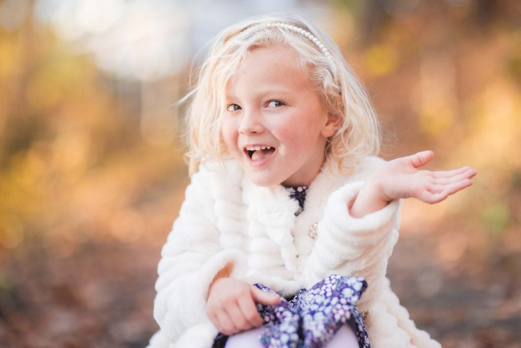 A joyful young girl with blonde hair, wearing a white fluffy jacket and a headband, laughing and gesturing with one hand in a sunlit autumnal forest. Perfect for family portraits.