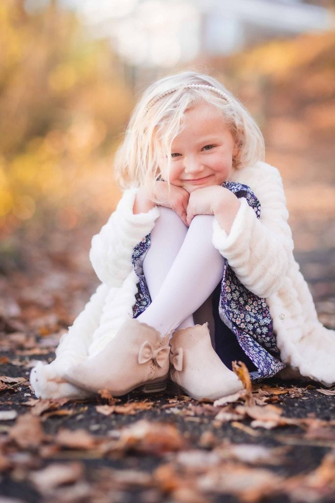 Young girl with blonde hair smiling, sitting on autumn leaves while wearing a white coat, patterned dress, and beige boots during a family portraits session.