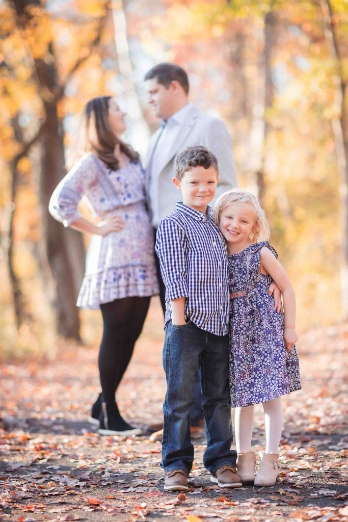 A family poses for family portraits in an autumn forest; two young children smile in the foreground while their parents, slightly out of focus, stand behind them.