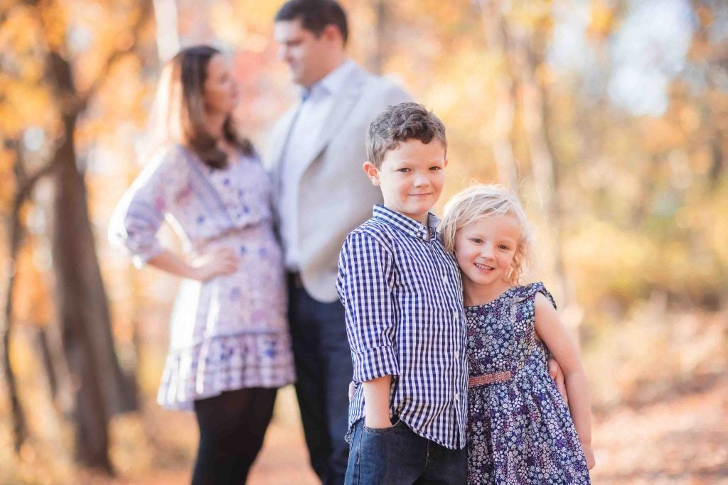 A young boy and girl pose smiling in the foreground with blurred parents standing in the background amidst autumn foliage for a family portrait.