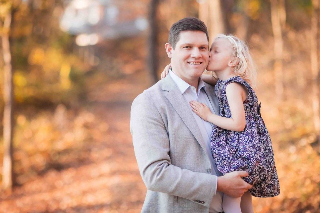 A young girl kisses her father on the cheek in a sunlit autumn forest, capturing a moment perfect for Family Portraits.