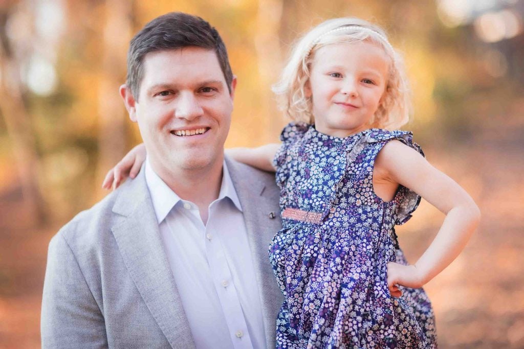 A man in a gray blazer and a young girl in a floral dress posing together with a softly blurred autumnal background for family portraits.