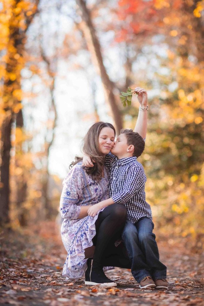 A young boy kisses his mother on the cheek while holding a leaf above her head, both seated in a sunlit autumn forest, perfect for family portraits.