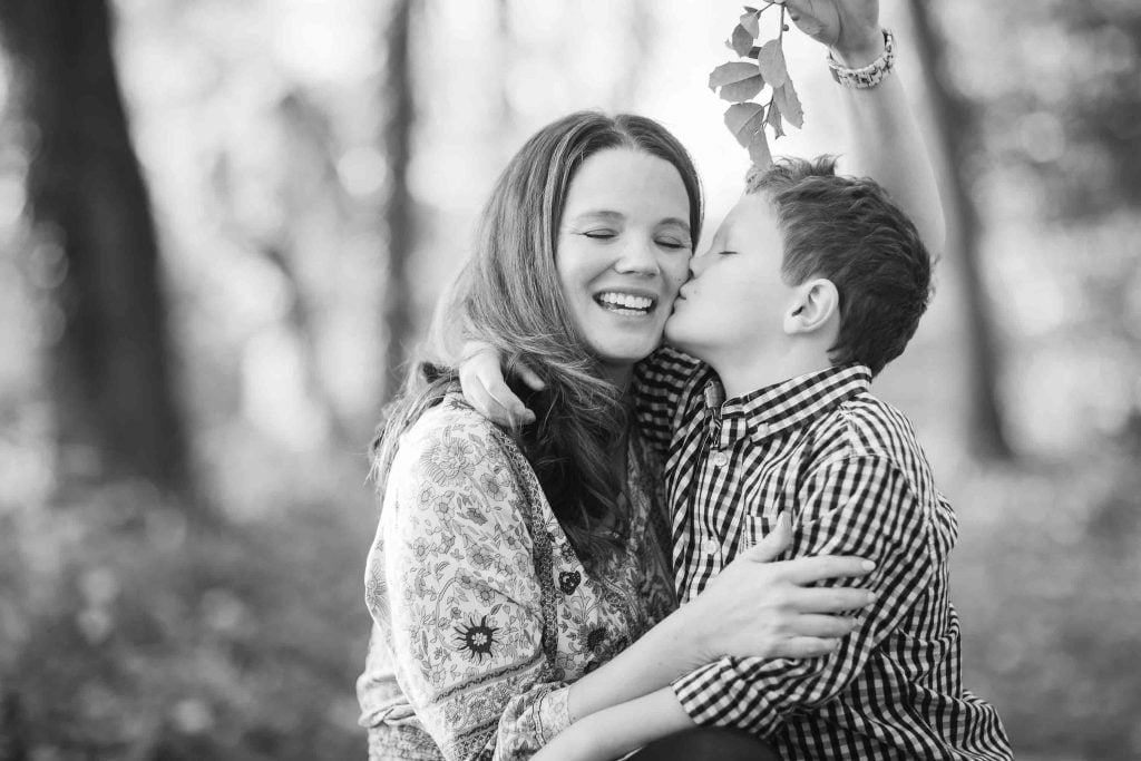 Black and white Family Portraits image of a smiling woman and a boy in a plaid shirt, with the boy kissing the woman on the cheek in a forest setting.