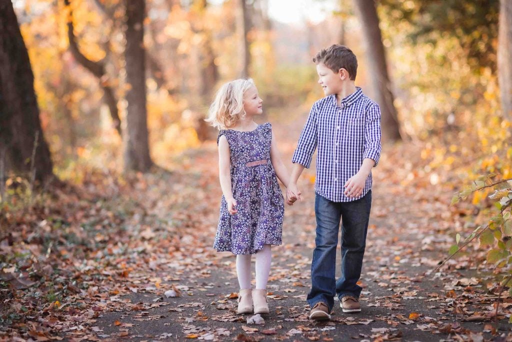 Two children, a boy and a girl, holding hands and looking at each other on a forest path surrounded by autumn leaves in Family Portraits.