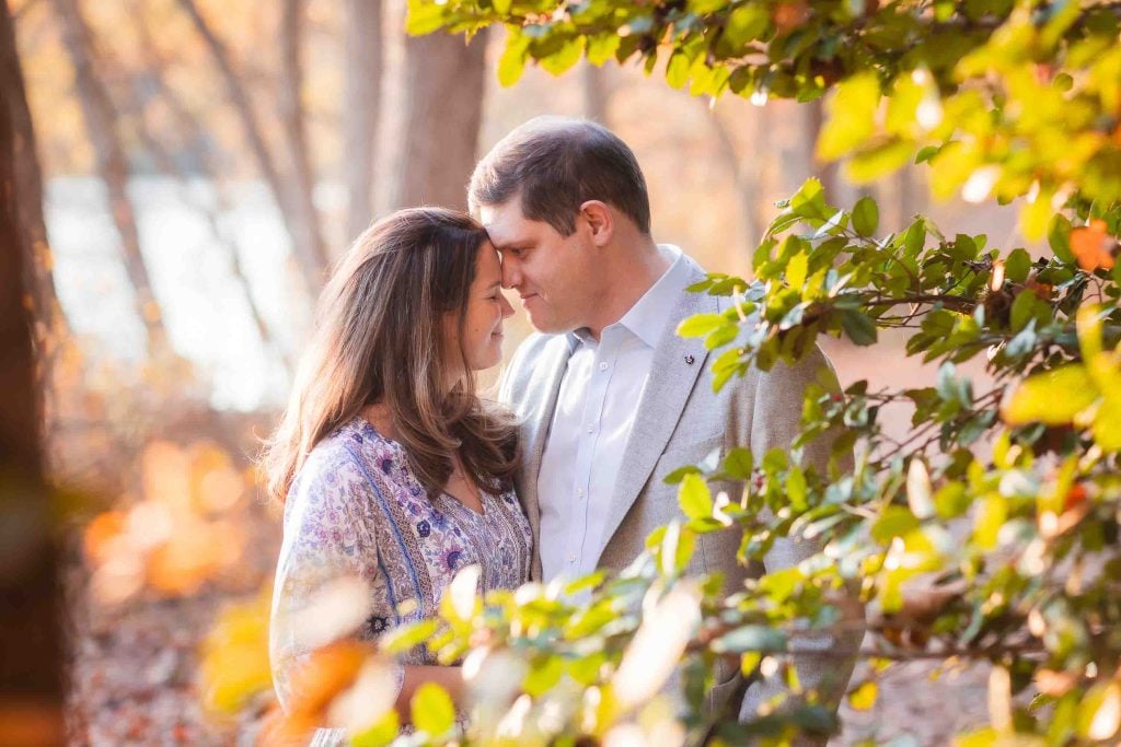 A family portrait of a couple affectionately touching foreheads, surrounded by autumn leaves in a forest, bathed in soft sunlight.