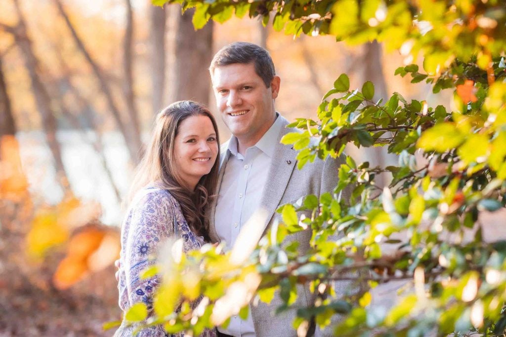 A smiling couple standing together among colorful autumn foliage in a family portrait, with soft sunlight illuminating the scene.