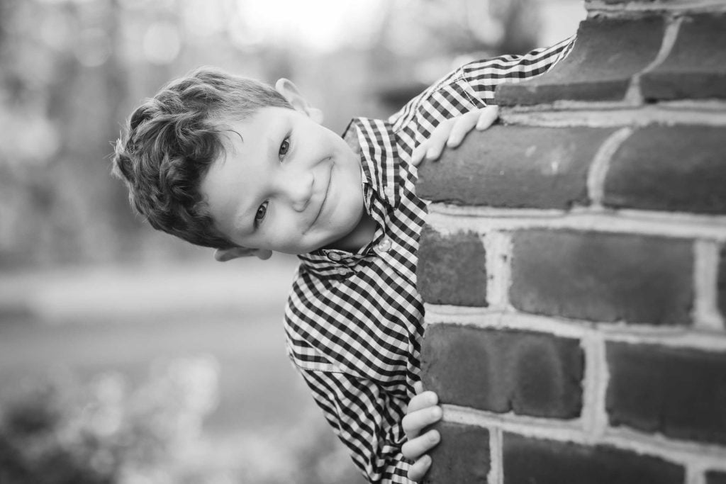 Young boy leaning playfully around a brick wall, smiling at the camera in a black and white family portrait photograph.