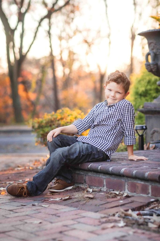 A young boy in a plaid shirt and jeans sitting on a brick ledge, smiling slightly, with autumn leaves in the background, perfect for family portraits.