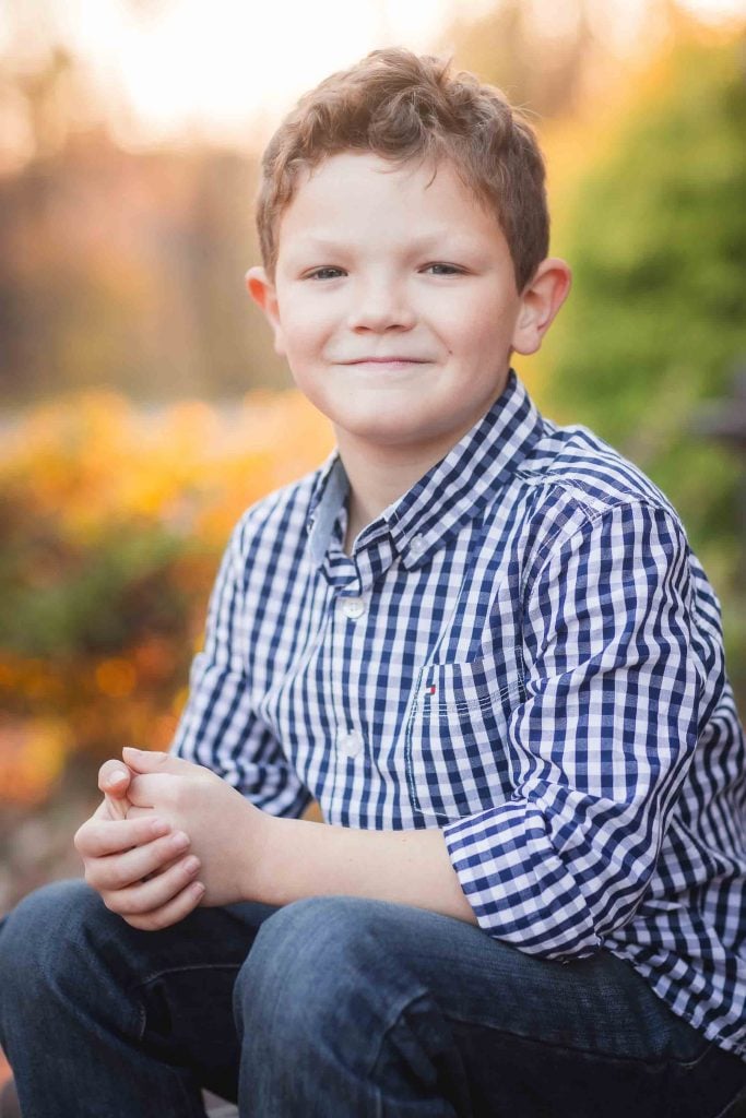 A young boy smiling at the camera, wearing a blue checkered shirt, seated outdoors with autumn leaves in the background, perfect for family portraits.