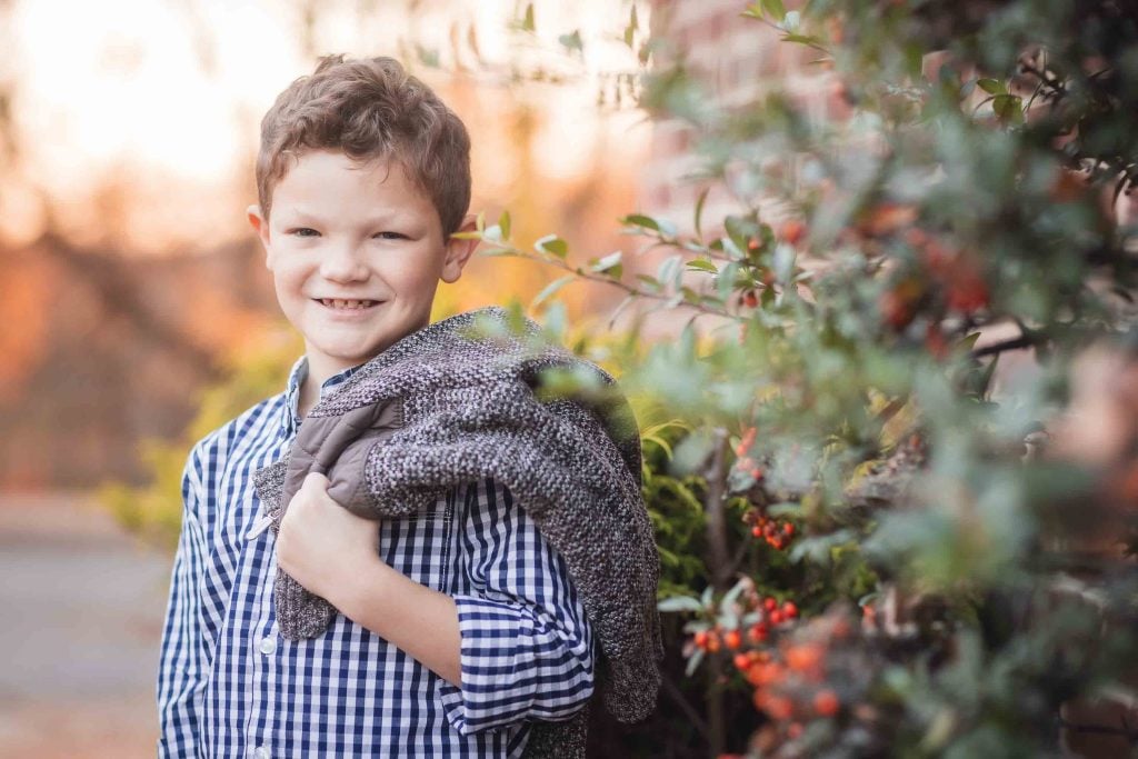 A young boy smiling at the camera, holding a gray sweater, standing next to a berry bush in a sunlit setting, perfect for family portraits.