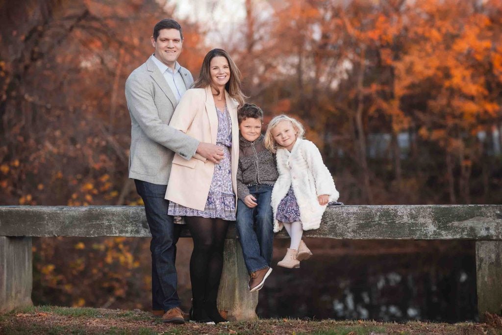 A family portrait of four posing happily on a wooden fence with autumn trees in the background, all smiling.