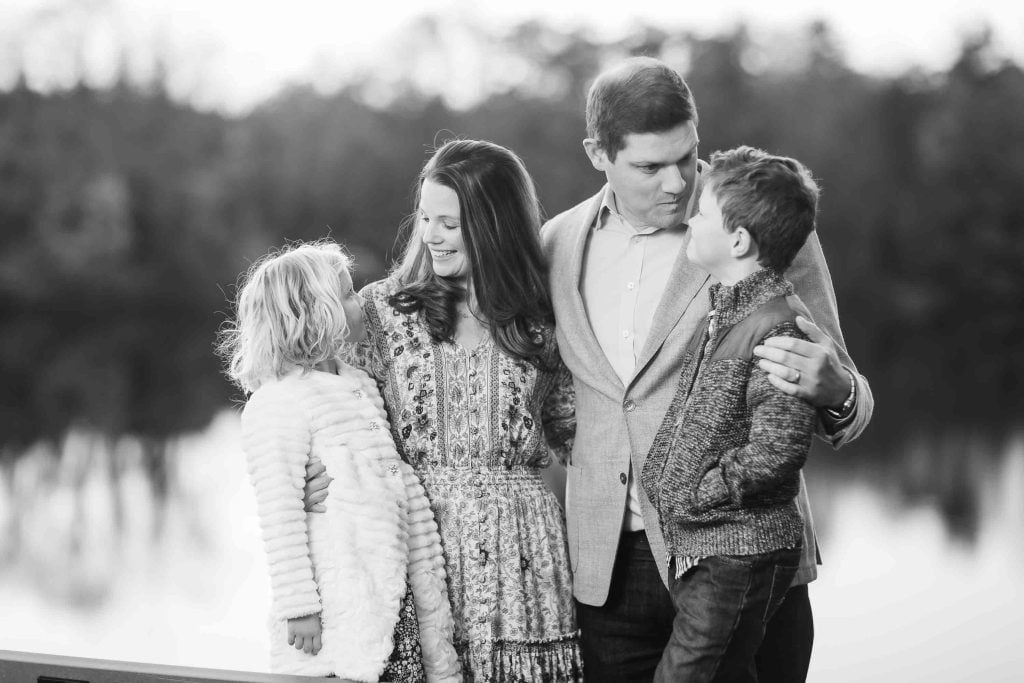 A black and white Family Portraits photo of four, with a man embracing two children while looking at a woman, all standing against a blurred natural background.