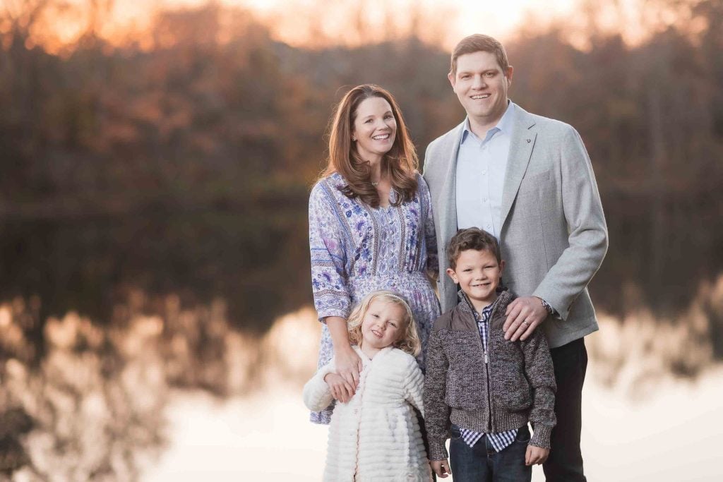 A family of four smiling by a lake at sunset, surrounded by autumncolored trees, perfect for Family Portraits.