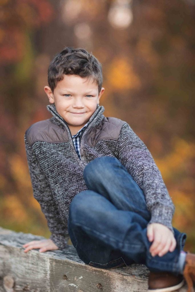 A young boy sitting on a wooden fence with colorful autumn leaves in the background, ideal for family portraits. He is wearing a sweater, jeans, and has a slight smile.