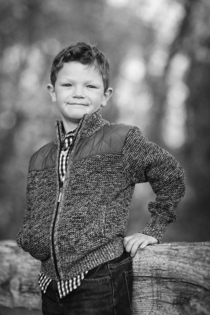 A young boy in a knitted jacket and checkered shirt leans on a wooden fence, smiling, in a blurred forest background. This black and white photo is an example of family portraits.