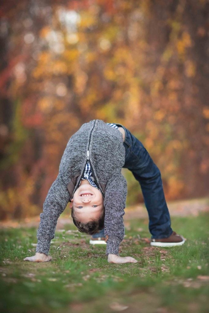 A young boy in a gray sweater performing a backbend on a grassy area with colorful autumn leaves in the background, ideal for family portraits.