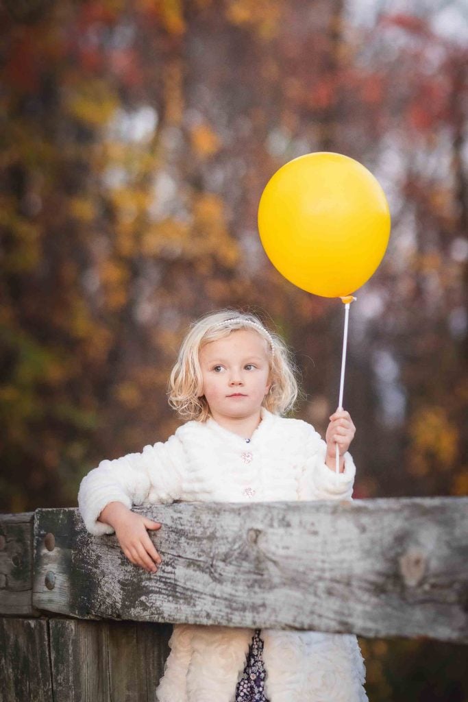 Young child holding a yellow balloon while leaning on a wooden fence with autumn leaves in the background, perfect for Family Portraits.