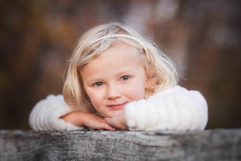 A young child with blonde hair, resting their chin on their hands on a wooden ledge, looking directly at the camera with a soft background in this family portrait.