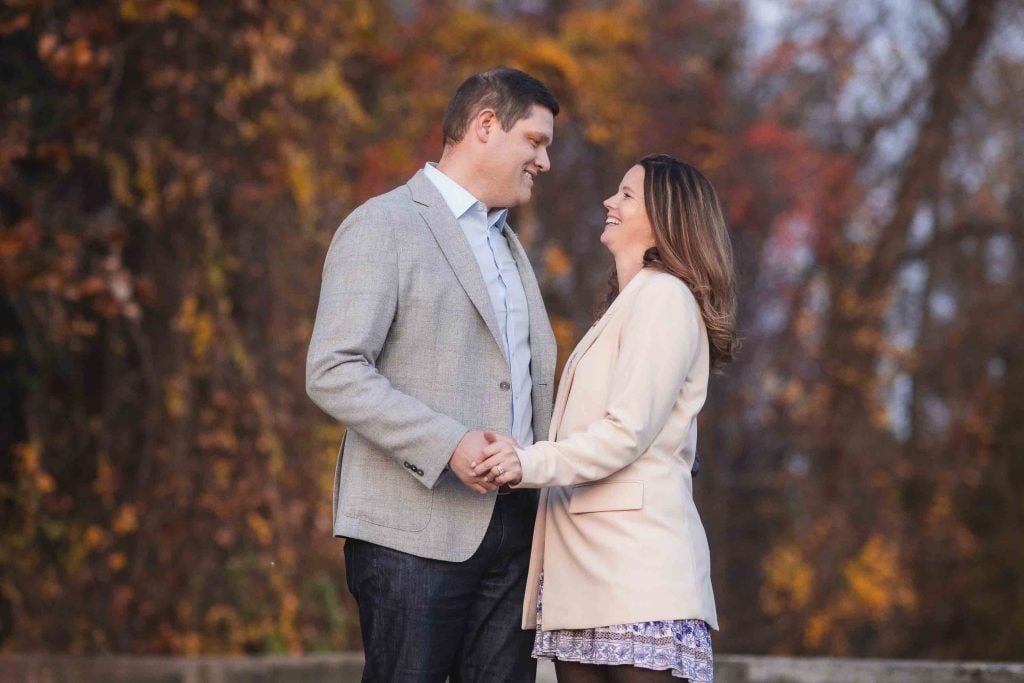 A couple smiling at each other, holding hands in an autumn park with colorful trees in the background, perfect for family portraits.