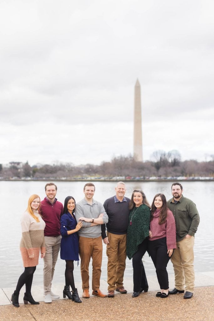 A family of seven stands together in front of the Washington Monument, with the overcast sky and water creating a beautiful backdrop reminiscent of classic portraits.