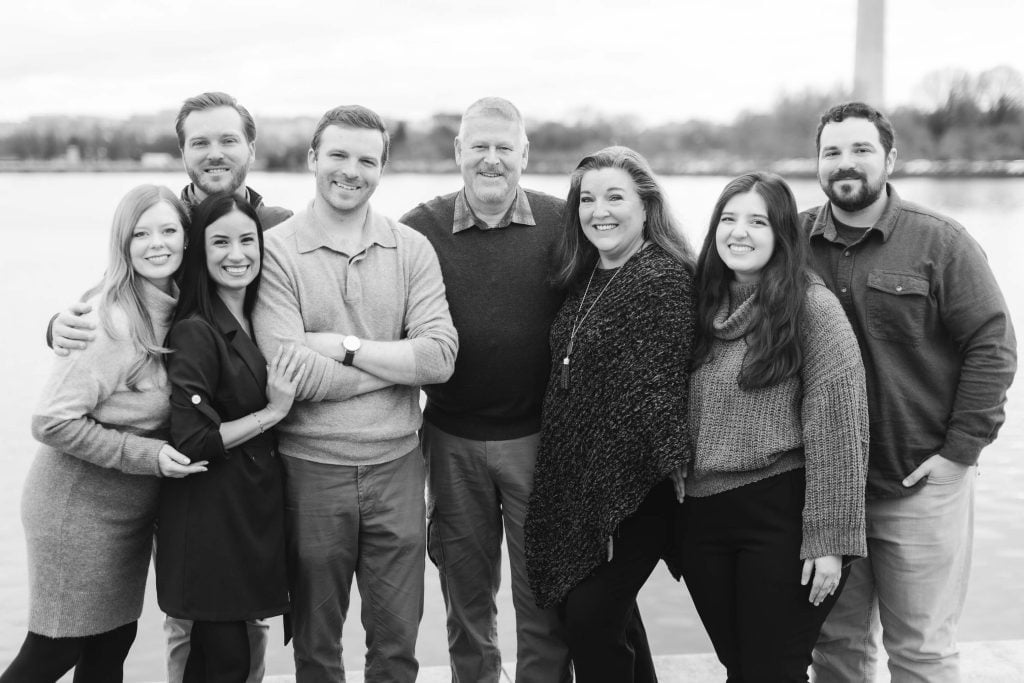 A group of eight people standing in two rows by a body of water, with the Jefferson Memorial towering in the background. Everyone is smiling and wearing casual winter clothing. The blackandwhite image has a timeless quality, almost like a family portrait from another era.