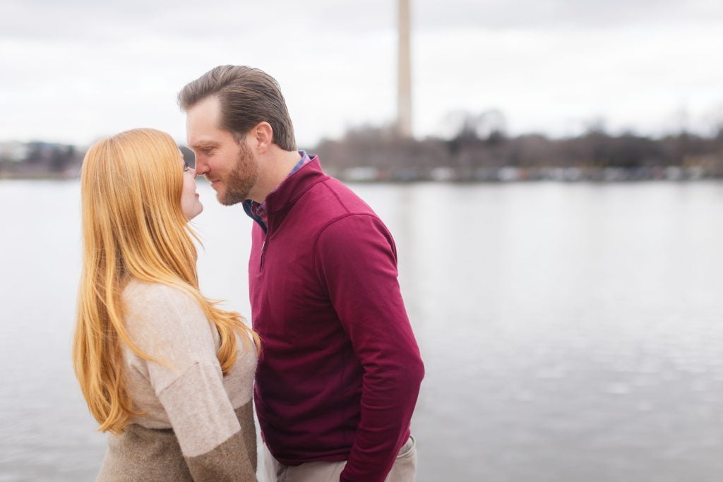 A man and a woman face each other closely near a body of water with the Washington Monument in the background, their intimate moment reminiscent of timeless family portraits.