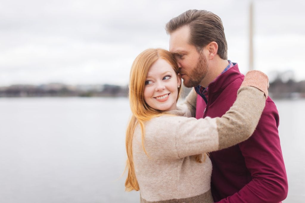 A man and woman embrace near a body of water with the Jefferson Memorial visible in the background. The woman smiles while the man kisses her on the forehead, creating a perfect moment reminiscent of timeless family portraits.