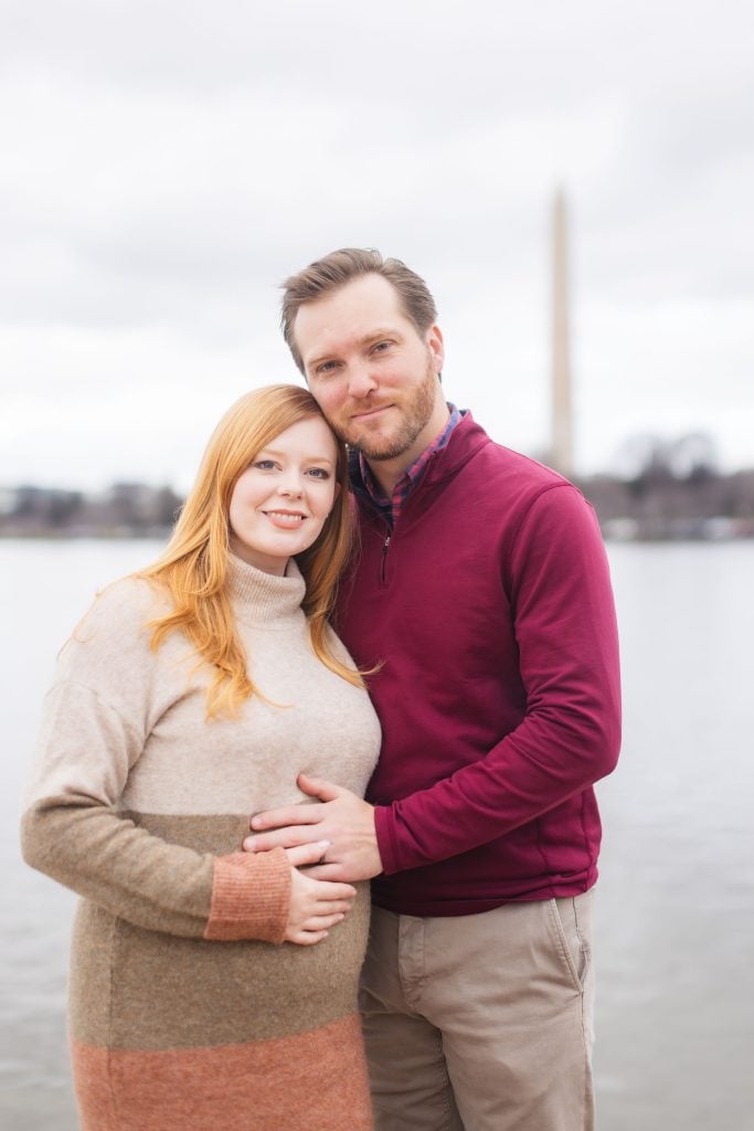 A couple stands close together by a body of water, with the man gently placing his hand on the woman's pregnant belly. The Washington Monument and Jefferson Memorial are visible in the background, capturing a serene moment in their family portraits.