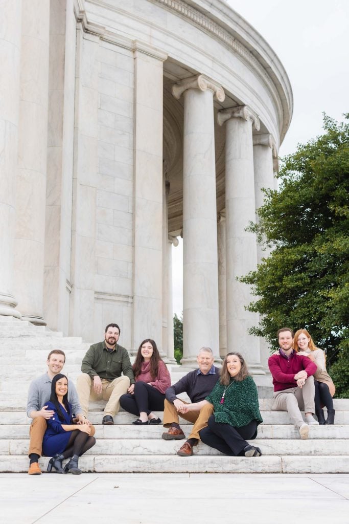 A family of seven people sits on the steps in front of a marble building with columns, posing for a photo. There's a green tree to the right, and it looks like they are at the Jefferson Memorial.