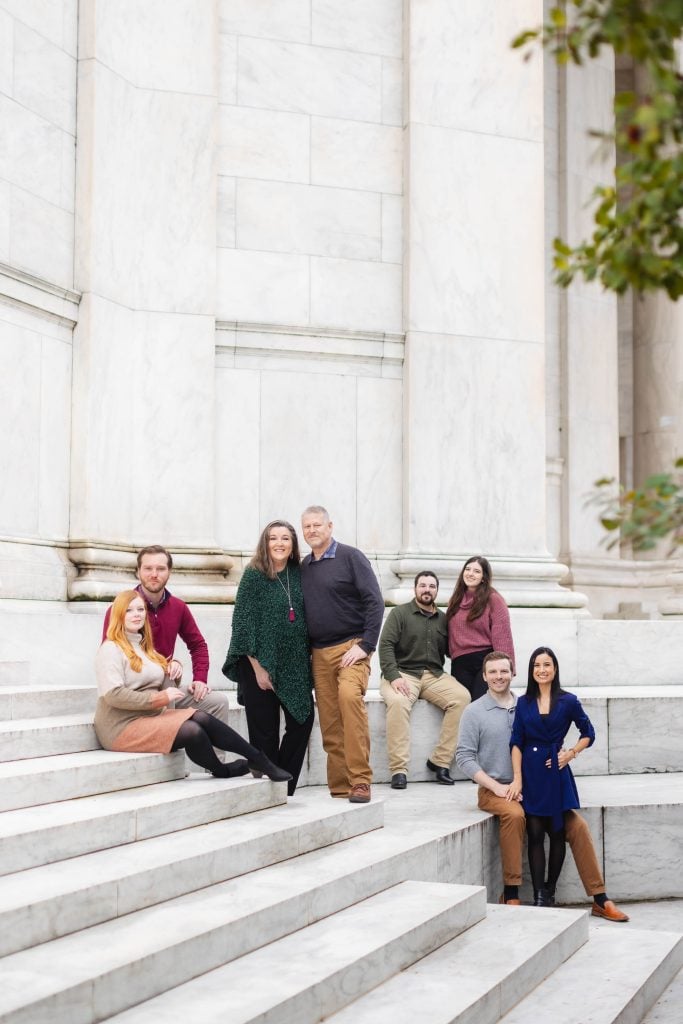 A family of seven poses on the steps of a marble building, reminiscent of the Jefferson Memorial. Some members are standing while others are seated, arranged in a casual formation. Arching columns loom gracefully in the background, creating an elegant setting for portraits.