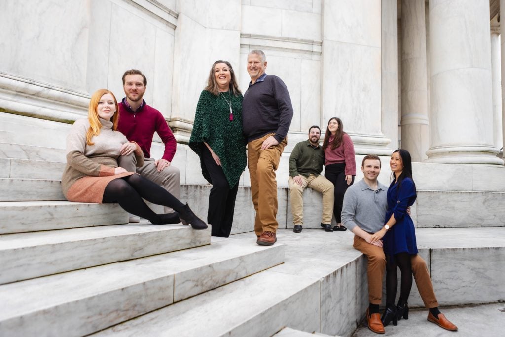 A group of seven people pose on the marble steps of the Jefferson Memorial with large pillars in the background. Four are seated, three are standing, all dressed in casual to semiformal attire—capturing a timeless family portrait.