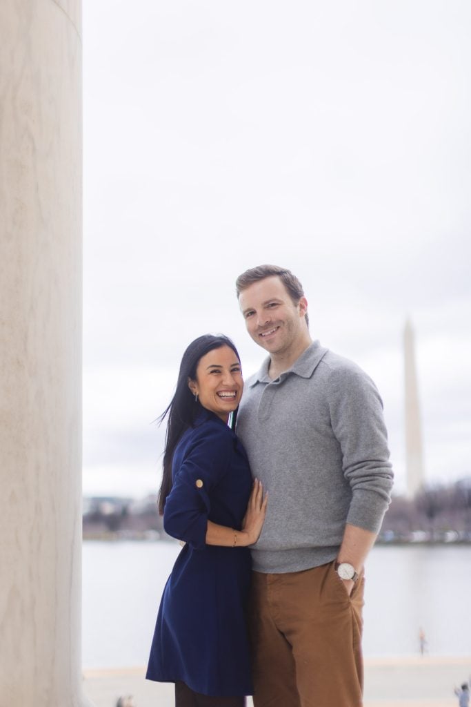 A woman and a man stand closely together, smiling, in front of a tall white column at the Jefferson Memorial, with the Washington Monument visible in the background.