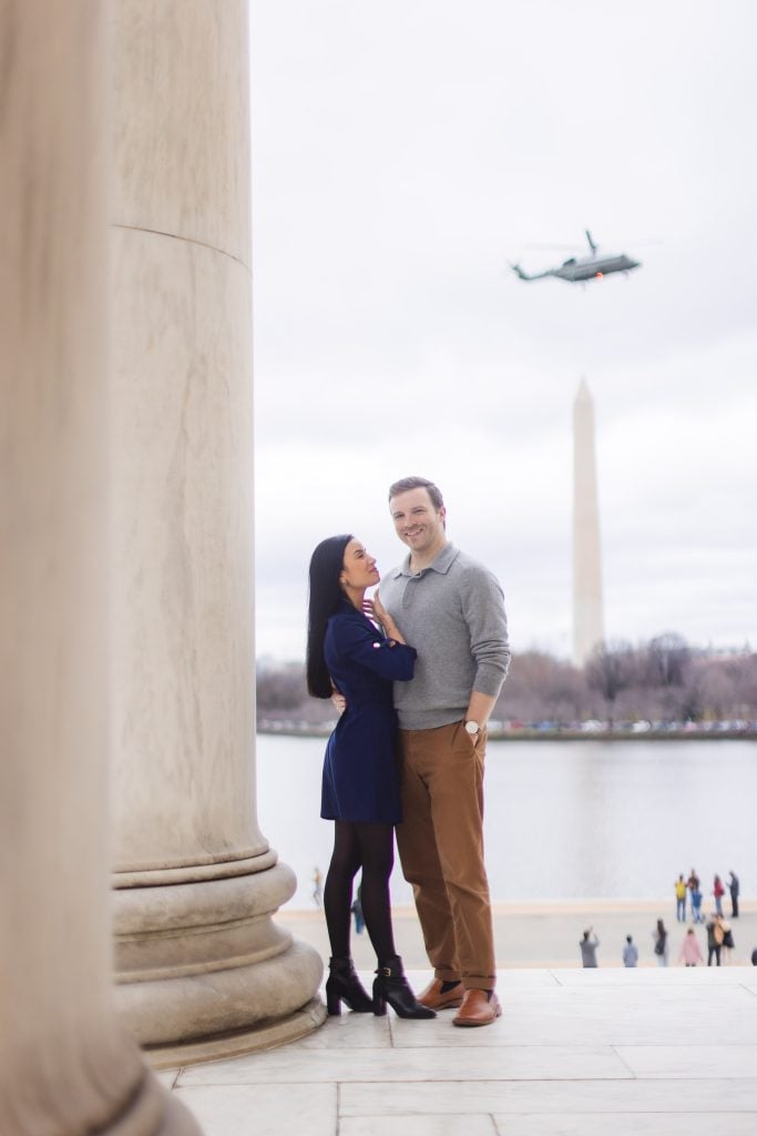 A couple poses by a column with the Washington Monument in the background and a helicopter flying above, capturing a cherished moment akin to family portraits near the Jefferson Memorial.