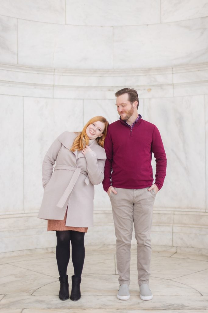 A woman and a man stand close together, smiling and dressed warmly in coats, against a white marble wall at the Jefferson Memorial. The woman is leaning her head on the man's shoulder, creating a perfect family portrait.