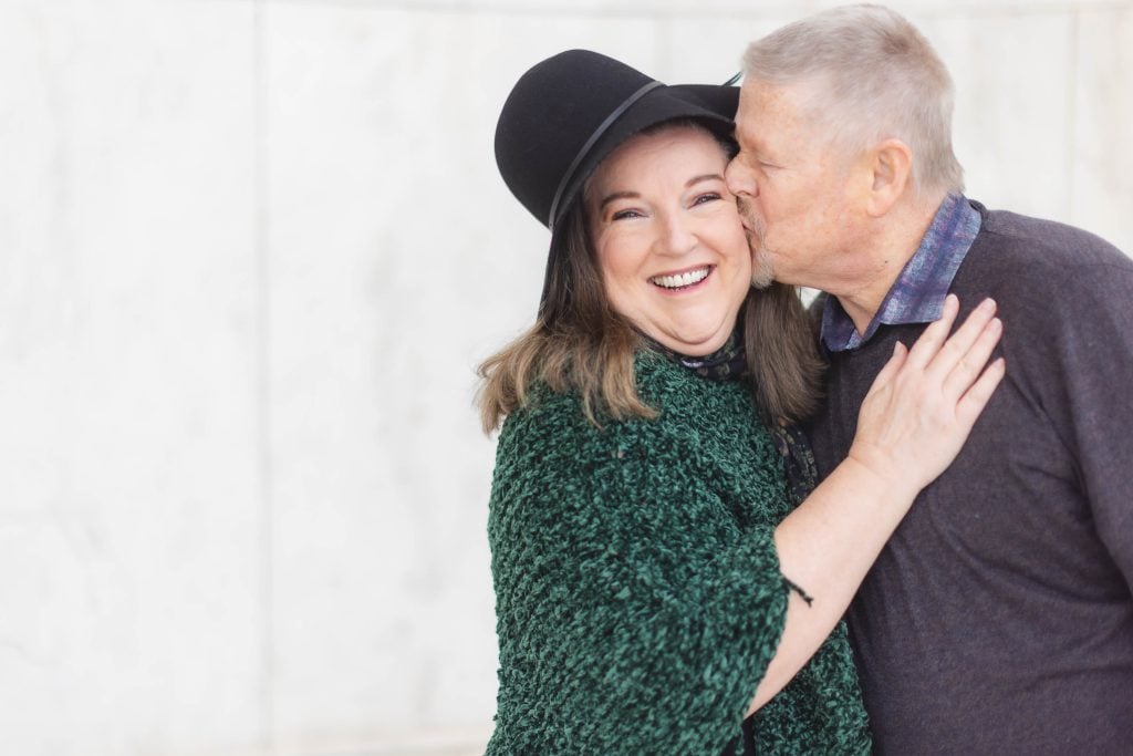 A smiling woman wearing a green shawl and black hat is being kissed on the cheek by an older man in a dark sweater. They stand in front of a lightcolored, slightly textured background, evoking the feel of cherished family portraits.