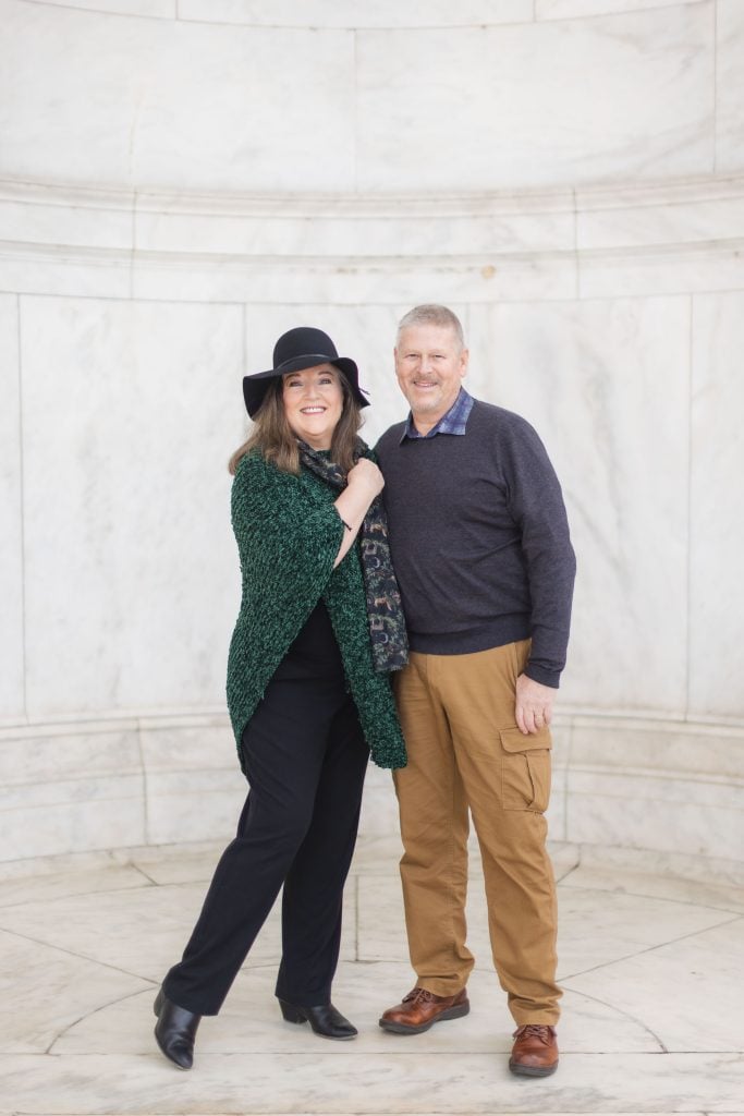 A woman in a green shawl and black hat stands next to a man in a gray sweater and beige pants. They both smile and pose for portraits in front of a white marbled backdrop, reminiscent of the Jefferson Memorial.