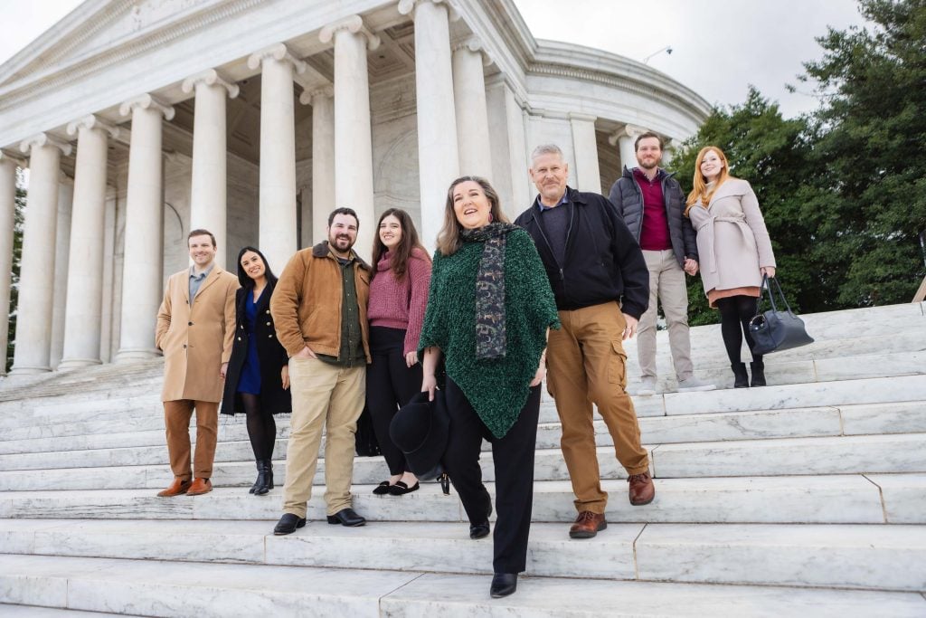 A family, dressed in winter clothing, stands on the steps of a neoclassical building with columns, smiling and looking towards the camera. The scene resembles classic portraits taken at iconic locations like the Jefferson Memorial.