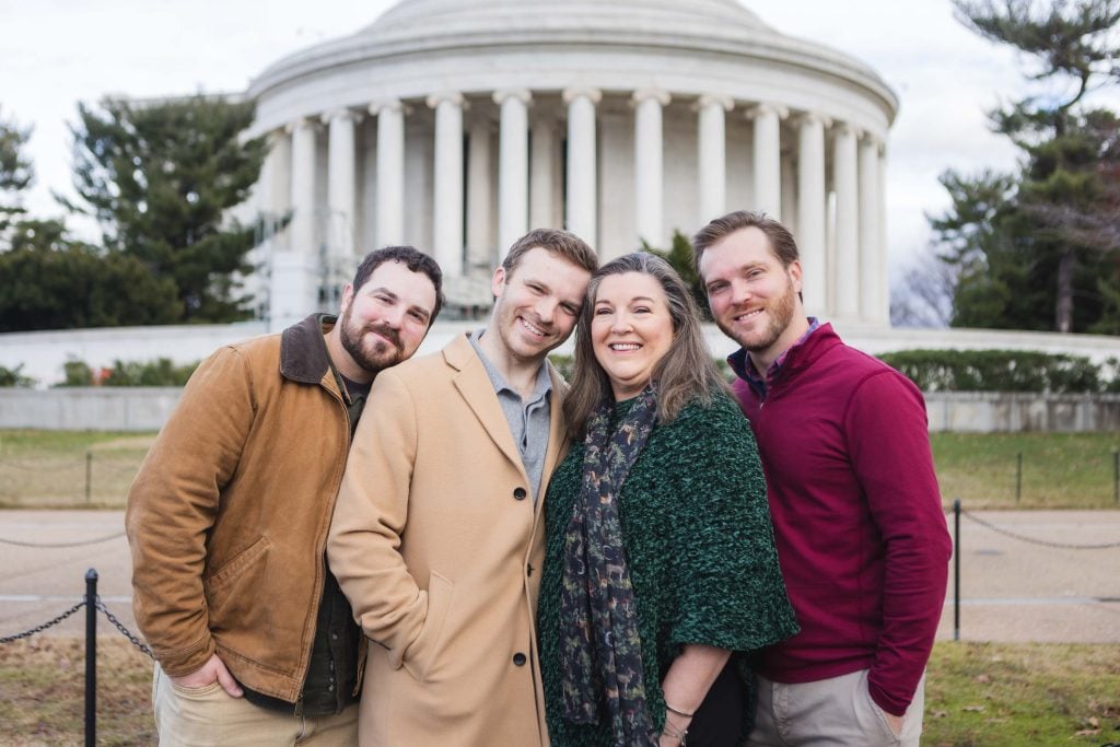 Four smiling adults stand closely together in front of the Jefferson Memorial, a white, columned building with a domed roof. They are outdoors, and the weather appears to be cool—a perfect setting for family portraits.