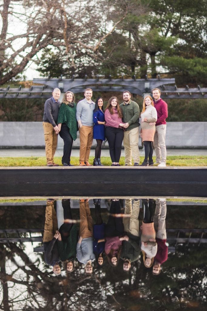 A group of seven adults, posed outdoors by a reflecting pool near the Jefferson Memorial. The background includes trees, a wall, and clear skies. Their reflection is visible in the water, capturing the essence of family portraits in this serene setting.